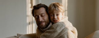 A bearded man and a blond child share a warm hug on a sofa, smiling in soft natural light from a nearby window.
