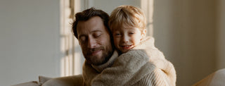 A bearded man and a blond child share a warm hug on a sofa, smiling in soft natural light from a nearby window.