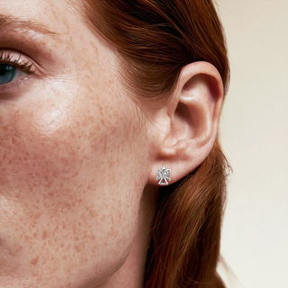A close-up of a woman's ear wearing a small silver angel stud earring, with her red hair and freckled skin visible against a white background.