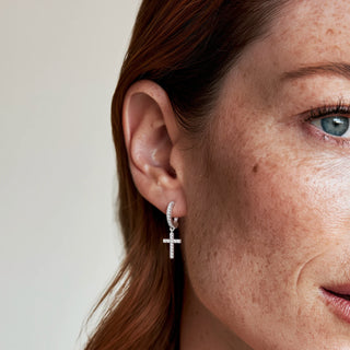 A close-up of a woman's ear wearingsilver and zirconia cross earrings, against a plain light background.