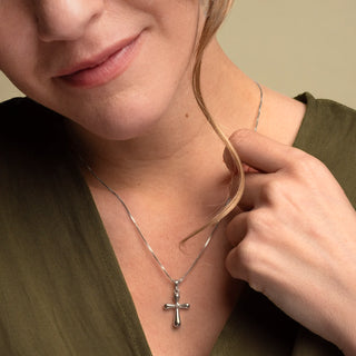 A close-up lifestyle shot of a woman wearing a delicate white gold cross pendant necklace on a fine chain, paired with an olive green top against a soft beige background.
