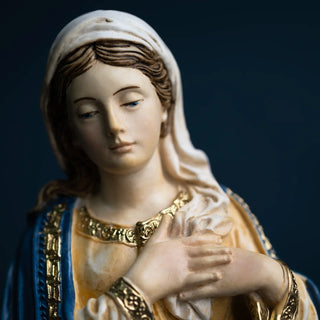 A close-up of a religious figurine depicting the Virgin Mary with a white head covering, gold and beige robes, and a blue mantle with gold trim, set against a dark blue background.