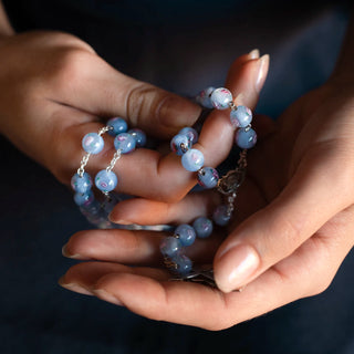 Hands holding a Light blue Murano glass rosary bead against a dark background