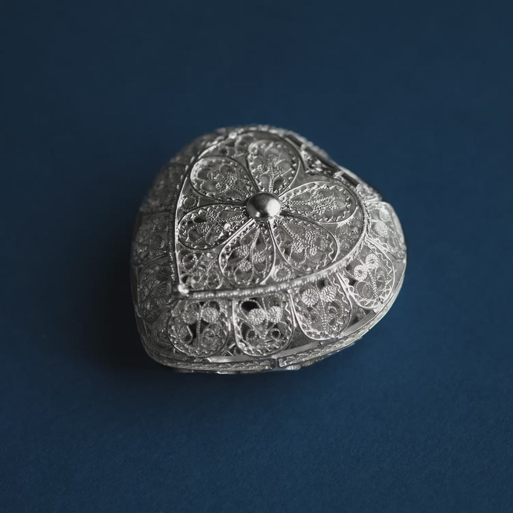 A silver filigree heart-shaped religious keepsake box with its lid open, revealing a matching silver rosary with beads and a cross rests in front on a dark blue background.