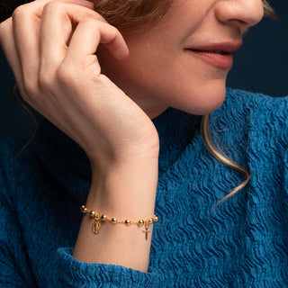A close-up of a woman wearing a delicate gold rosary bracelet with a small cross charm and a miraculous medal, paired with a textured blue sweater.