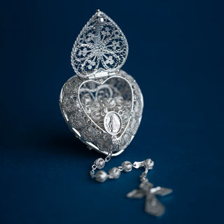 A silver filigree heart-shaped religious keepsake box with its lid open, revealing a matching silver rosary with beads and a cross rests in front on a dark blue background.